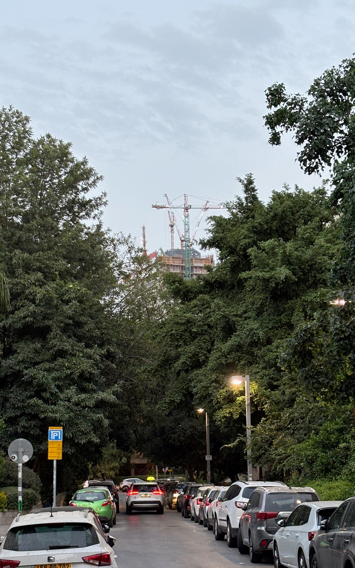 Street scene with cars parked along a tree-lined road, construction crane in the background.