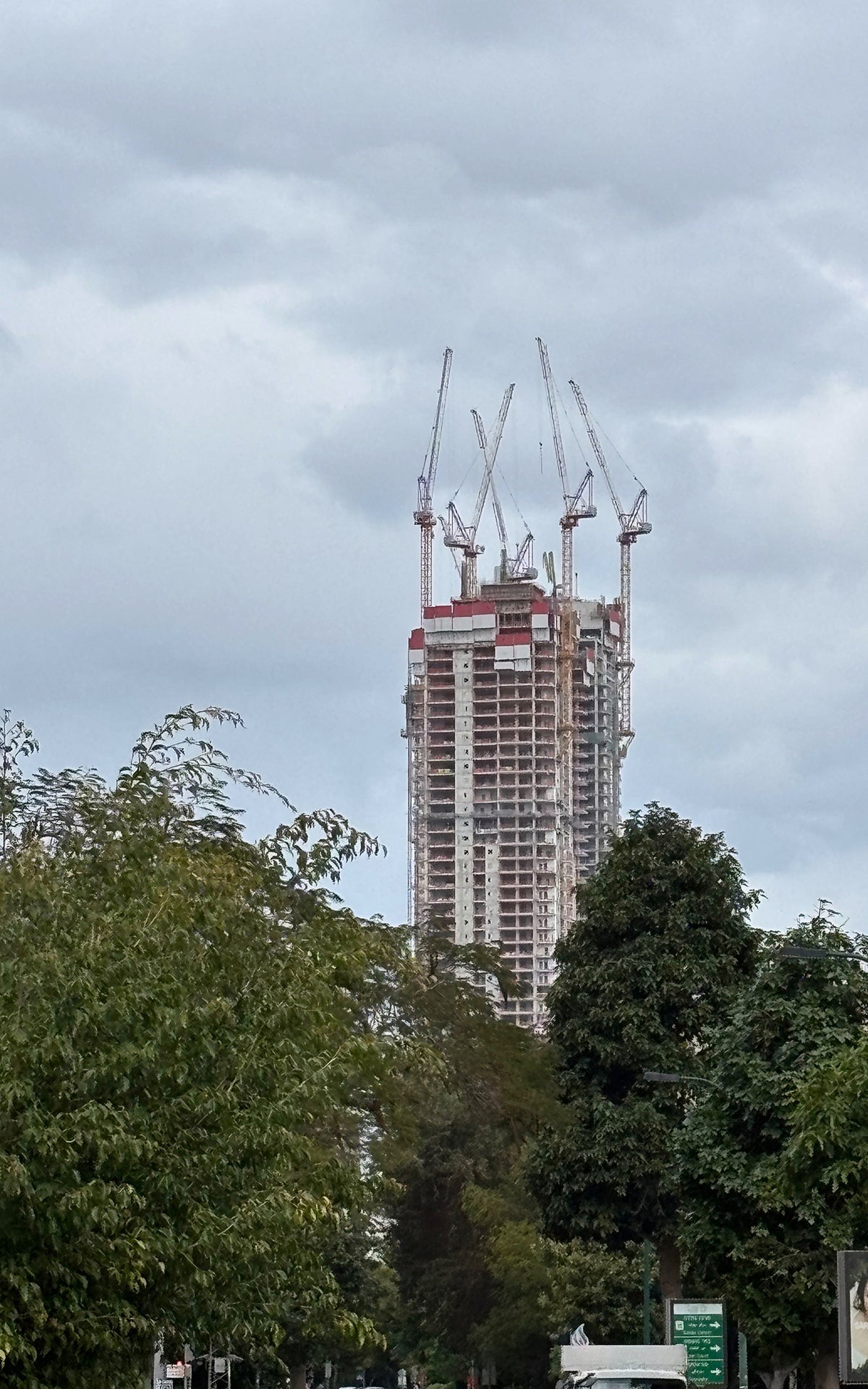 Quiet street in TLV center with tall building under construction with cranes against a cloudy sky appearing above.